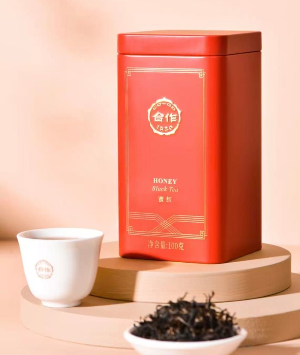 A red tin of CO-OP 1950 Honey Black Tea beside a white porcelain cup and a small dish of dry leaves